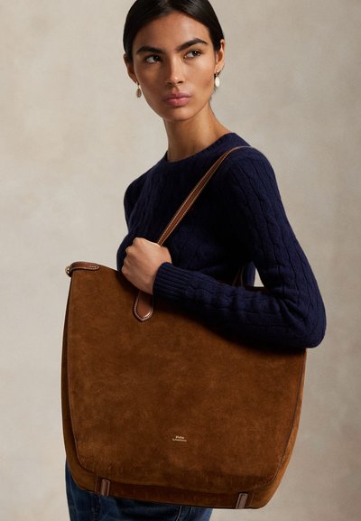 Femme aux cheveux foncés portant un pull bleu marine tient un grand sac cabas en daim marron avec des bandoulières en cuir sur l'épaule.
