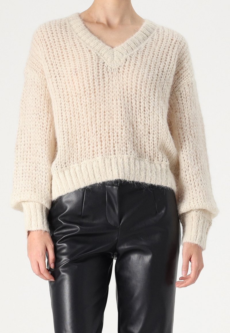 Personne portant un pull crème à col en V ample en maille et un pantalon taille haute en similicuir noir, debout les mains détendues.