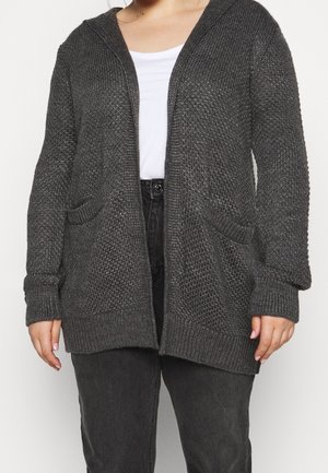 Persona con cárdigan de punto gris oscuro con bolsillos frontales sobre una camiseta blanca y pantalones negros, de pie ante un fondo claro y liso.