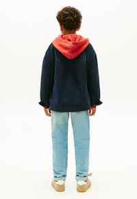 Dżinsowa kurtka w ciemnoniebieskim kolorze, nałożona na czerwoną bluzę z kapturem. Jasnoniebieskie dżinsy z lekkim przetarciem, połączone z beżowymi trampkami. Prosty design, bez wzorów.