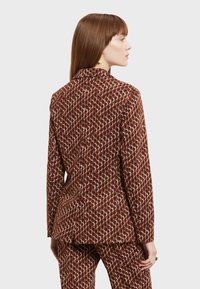 Ruskean kuviollinen bleiseri, jossa on muotoiltu istuvuus, hienovarainen geometristen muotojen kuviointi kermaisena ja tummanpunaisena, sileä kangas ja lovellinen kaulus.