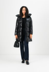 Zwarte pufferjack met glanzende afwerking, capuchon en ritsluiting. Gecombineerd met marineblauwe jeans en zwarte instappers, met een kleine zwarte tas.