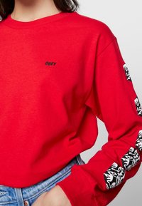 Röd långärmad skjorta i bomull med en svart "OBEY"-logotyp på bröstet och ett grafiskt svartvitt mönster på ärmarna.