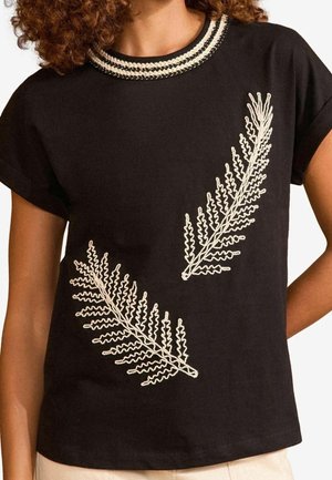 Vrouw draagt een zwart top met korte mouwen met beige varenbladborduurwerk en een gevlochten beige-zwart halslijn.