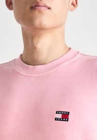 Sudadera de color rosa claro con cuello redondo, que presenta un pequeño parche de logo "TOMMY JEANS" en negro y rojo en la parte izquierda del pecho. Textura suave.