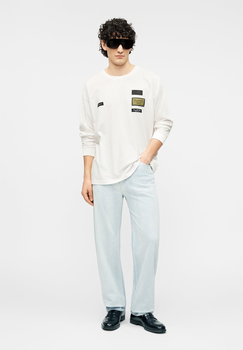 Hvid langærmet skjorte med stoflapper, kombineret med lyseblå jeans og sorte sko. Modellen bærer store sorte solbriller.