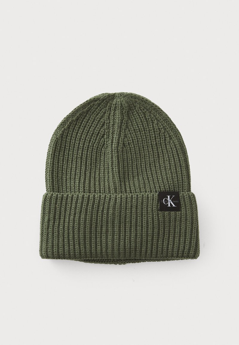 Olijfgroene gebreide beanie gemaakt van ribgebreid materiaal, met een omgeslagen rand en een zwart logo-embleem aan de zijkant.