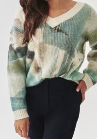 Frau trägt einen weichen, mehrfarbigen Strickpullover in Blau-, Grün- und Beigetönen, kombiniert mit hoch taillierten schwarzen Hosen.