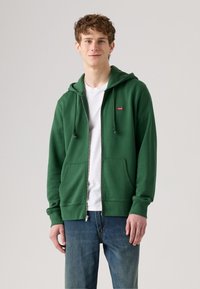 Felpa verde con zip realizzata in tessuto morbido, dotata di cappuccio regolabile con cordino, tasche frontali e un piccolo logo rosso sul lato sinistro del petto.