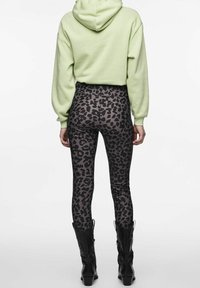 Osoba oblečená v světle zelené mikině s kapucí, černých a šedých legínách s leopardím vzorem a černých kozačkách ke kolenům, stojící zády k objektivu na bílém pozadí.
