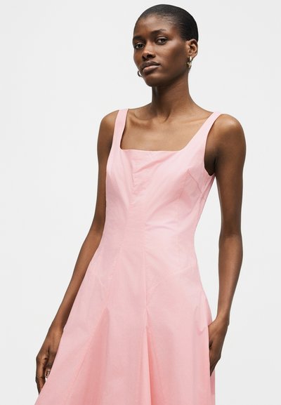 Γυναίκα που φοράει αμάνικο ανοιχτό ροζ φόρεμα με τετράγωνο ντεκολτέ, στέκεται μπροστά σε απλό φωτεινό γκρι φόντο.