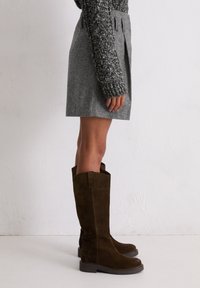 Jupe en mélange de laine grise avec des plis, associée à des bottes hautes en suède marron avec une semelle épaisse et un détail en onglet à l'arrière.