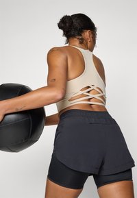 Beige sport-BH med kryssryggdesign, kombinert med svarte lagdelte shorts. Antrekket har en jevn tekstur og en tettsittende silhuett.