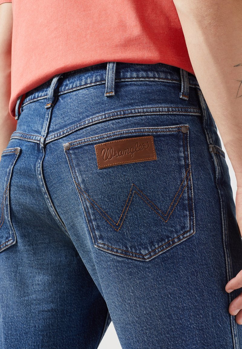 Vaqueros de mezclilla azules con un parche de cuero marrón etiquetado como "Wrangler", que presenta costuras naranjas contrastantes y un diseño de zigzag distintivo en los bolsillos traseros.