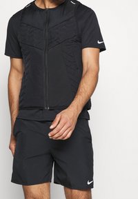 Schwarz sportlicher Zip-Up-Weste mit strukturierten Einsätzen und kurzen Ärmeln, kombiniert mit schwarzen Shorts. Nike-Logo auf der Weste und den Shorts.