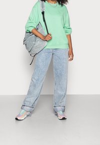 Sudadera verde menta, jeans holgados azul claro, zapatillas iridiscentes y una mochila gris a rayas, mostrando un estilo relajado y casual.