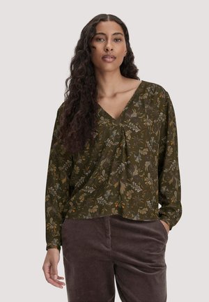 Vrouw met lang krullend haar die een groene gebloemde V-hals blouse en donkerbruine corduroy broek draagt, staand met één hand in de zak.