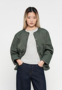 Jeune femme avec de longs cheveux ombrés portant une veste matelassée vert foncé, un t-shirt gris clair et un jean bleu foncé, les mains dans les poches.