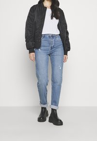 Giacca bomber nera sopra una maglietta bianca strutturata, abbinata a jeans chiari strappati e stivali alla caviglia neri e spessi. Design semplice e aderente.