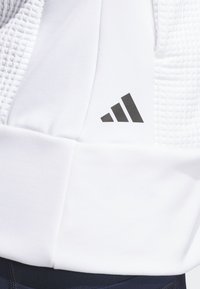 Detailní záběr na bílou texturovanou látku s černým logem Adidas vytištěným na hladké části poblíž tmavě modré látky níže.
