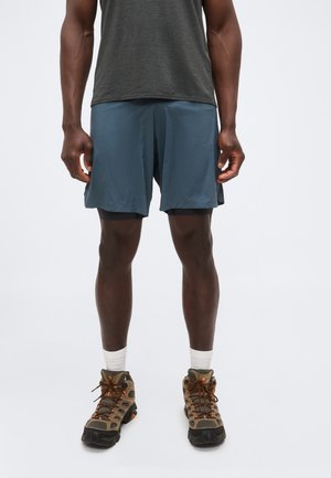 Férfi sötétszürke pólóban, kék sport rövidnadrágban, fekete kompressziós rövidnadrág fölött, fehér zokniban és barna túracipőben, egyenesen áll.