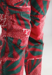 Rode stoffen broek met groene en witte abstracte bladerenpatronen. Voorzien van een voorzak, textuurmateriaal en gestikte details.