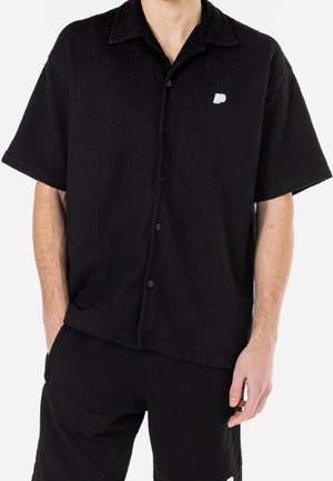 Mężczyzna ubrany w czarną, krótką, zapinaną na guziki teksturowaną koszulę i pasujące szorty, ręce częściowo w kieszeniach, na jednolitym tle.