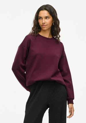 Sweatshirt bordeaux à col rond, épaules tombantes et manches longues, associé à un pantalon noir texturé. Tissu lisse, coupe décontractée.