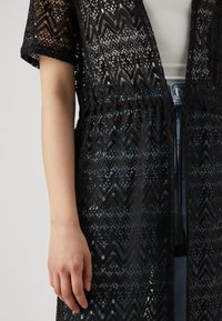 Svart blonde kimono med zigzag-mønster, korte ermer og snøring i midjen. Har en teksturert, åpen vev og dekorative duskdetaljer.