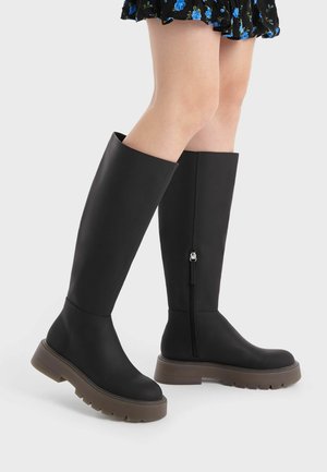 Mustat polvipituiset bootsit, jotka on valmistettu sileästä materiaalista, paksulla ruskealla pohjalla ja sivuvetoketjulla. Yksinkertainen, suoraviivainen malli ilman kuvioita.