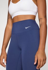 Marinblå, högmidjade leggings gjorda av elastiskt tyg med en mjuk yta, med en liten vit Nike-logotyp på midjebandet.