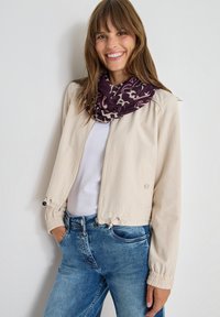Giacca beige accorciata con zip frontale, polsini elasticizzati e orlo con coulisse, abbinata a una sciarpa viola a motivi e a una maglietta bianca.