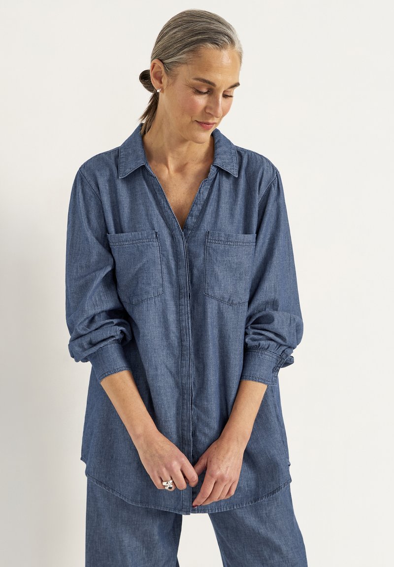 Chemise en denim avec un col, deux poches avant et des manches longues. Elle présente une coupe décontractée et un ourlet arrière plus long. Texture douce.