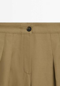 Pantalons khaki avec une fermeture à bouton à la taille, dotés de plis et d'une texture de tissu lisse. Aucun motif visible ni détails supplémentaires.