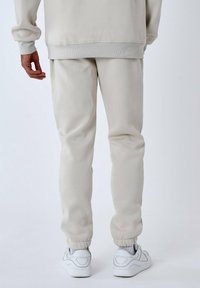 Pantalones de chándal beige con puños elásticos, textura suave, combinados con zapatillas blancas. La cinturilla es elástica y el corte es holgado.