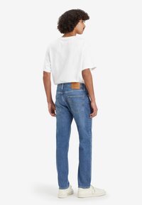 Jean en denim bleu clair avec une coupe droite, arborant une patch en cuir marron à l'arrière, porté avec un t-shirt blanc et des baskets blanches.