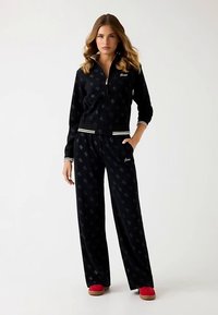 Black velour tracksuit with gold logo, zip-up jacket, and wide-leg pants. Features white and gold striped accents, soft texture, and all-over pattern.