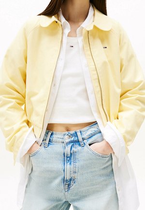 Person iført en gul lynlåstrøje over en hvid skjorte med knapper og en cropped hvid top, med lyseblå denimjeans og hænder i lommerne.