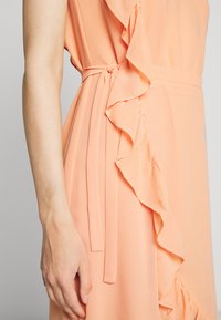 Robe pêche avec détail en volants sur le côté, nouée à la taille. Tissu doux et fluide avec des plis verticaux et une texture subtile.