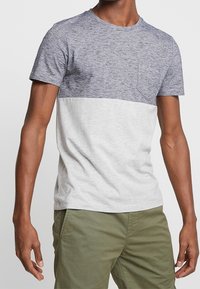 Hombre vistiendo una camiseta de dos tonos gris de manga corta con un bolsillo en el pecho y pantalones verde oliva contra un fondo sutil.