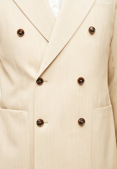 Μπεζ διπλόστενη σακάκι με έξι σκούρα καφέ κουμπιά και δύο εμπρόσθιες τσέπες, φορεμένο πάνω από ένα λευκό πουκάμισο.