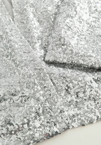 Zilverkleurige paillettenstof met een glanzend, reflecterend oppervlak. Kenmerkt zich door dicht bij elkaar geplaatste ronde pailletten die een textuurrijk, fonkelend uiterlijk creëren.