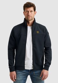 Dunkelmarineblaue Zip-Jacke mit einem strukturierten Oberteil, zwei seitlichen Taschen, gerippten Bündchen und Kragen, kombiniert mit einem weißen T-Shirt und blauen Jeans.