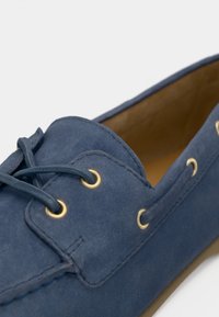 Κορδονάτο παπούτσι σκαφού σε ναυτικό μπλε δέρμα με χρυσά μεταλλικά νούμερα, επίπεδα κορδόνια και υφή φινιρίσματος. Το εσωτερικό είναι σε ταμπά χρώμα, τονίζοντας κλασικό σχέδιο.