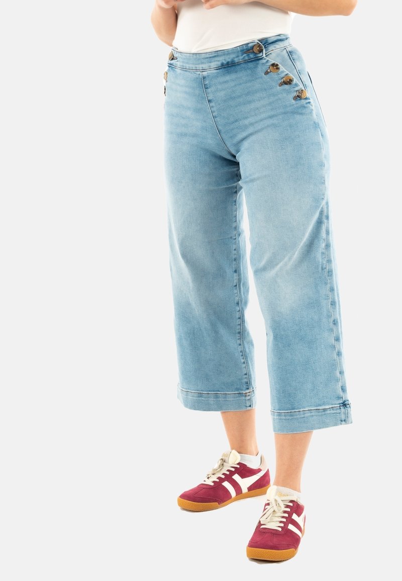 Personne portant un jean court large bleu clair avec des boutons sur le côté et des baskets rouges avec des bandes blanches.