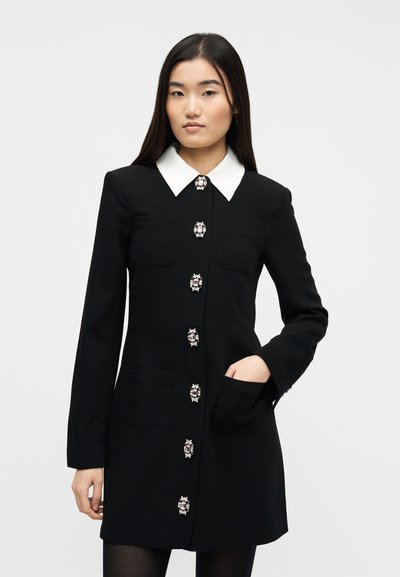 Νεαρή γυναίκα που φορά μαύρο φόρεμα με μακριά μανίκια, λευκό μυτερό γιακά και κουμπιά με κοσμήματα, με το ένα χέρι στην τσέπη, στέκεται σε απλό φόντο.