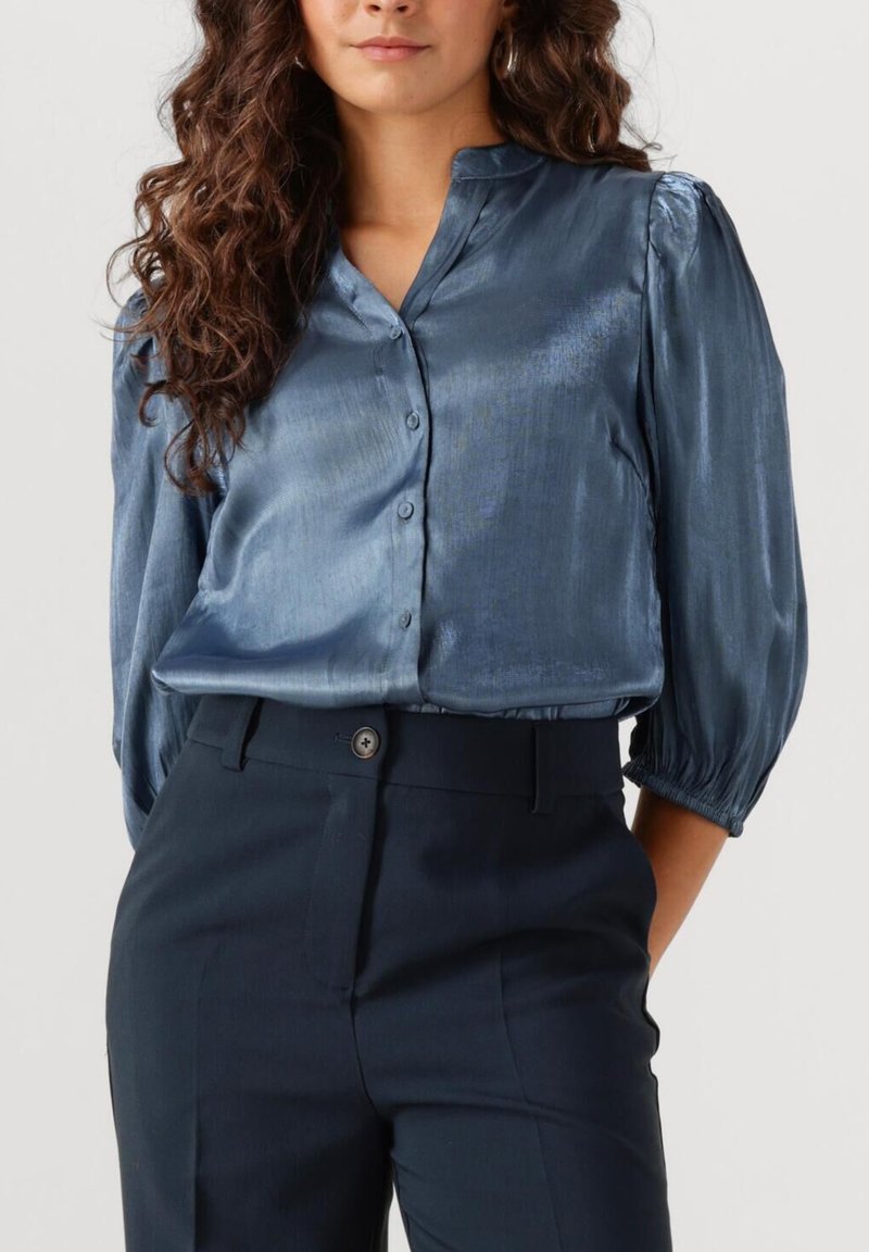 Blauwe satijnen blouse met een kraag, knoopsluiting aan de voorkant en korte pofmouwen. Gecombineerd met donkere, op maat gemaakte trousers met een knoop aan de voorkant.