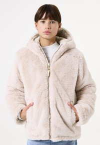 Chaqueta con capucha de felpa color beige, con cremallera en el frente, bolsillos laterales, textura suave y capucha forrada; presenta un corte relajado y hombros caídos.