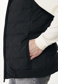 Ruka v kapse černé prošívané bundy oblečené přes bílé tričko s dlouhým rukávem a šedé kalhoty, zobrazeno z boku v úrovni pasu.