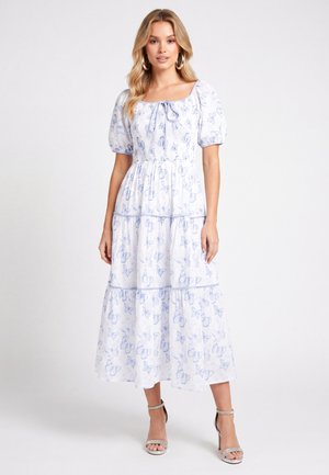 Woman wearing a white midi dress with blue butterfly print, puff sleeves, a square neckline with a bow, and silver high-heeled sandals.